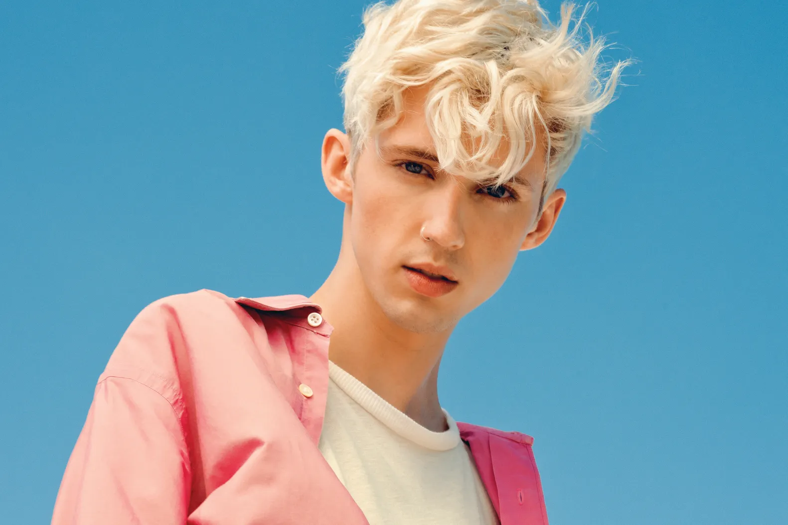 Portrait of a young man with platinum blond hair wearing a pink shirt over a white tee against a clear blue sky backdrop.