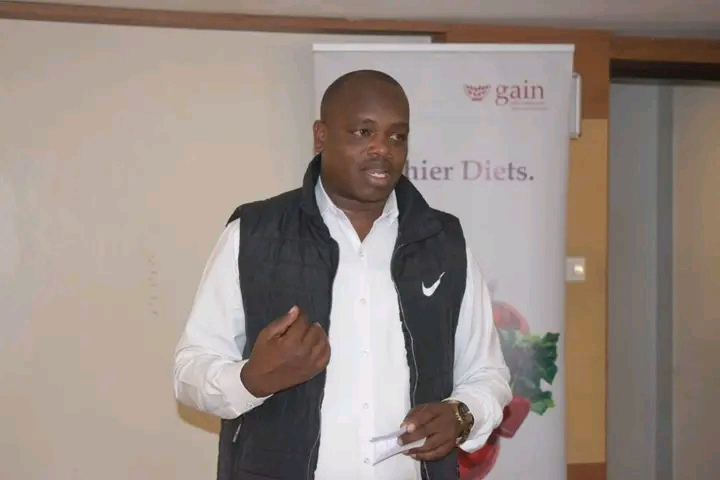 Man in white shirt and black puffer vest giving a talk indoors, holding papers in one hand.