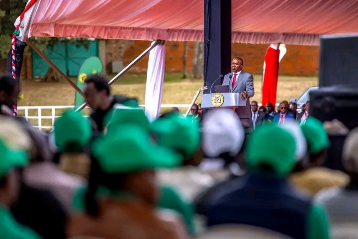 Man in a suit speaks at a lectern with a seal under an orange canopy, as an audience wearing green hats looks on.