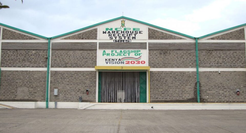 Front view of a brick warehouse with green trim and a large sign reading NCPB Warehouse Receipt System, Kenya Vision 2030 banner above the loading bay