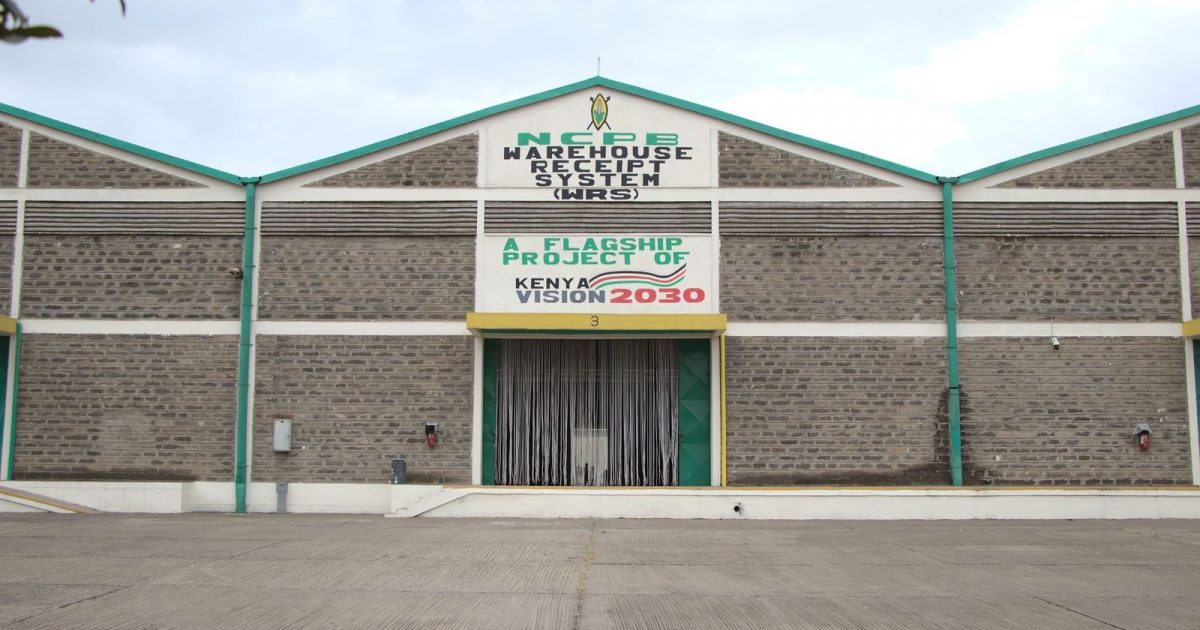 Front view of a brick warehouse with green trim and a large sign reading NCPB Warehouse Receipt System, Kenya Vision 2030 banner above the loading bay