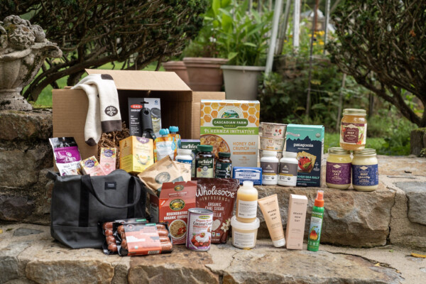 Assortment of groceries and personal care products arranged on a stone step outdoors, including boxes, jars, bottles, a tote bag, and a honeycomb-pattern gift box.