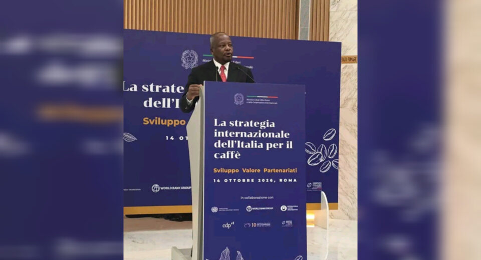 Man in a suit giving a speech at a conference, standing at a blue lectern with Italian coffee-themed banners behind him and on the podium.