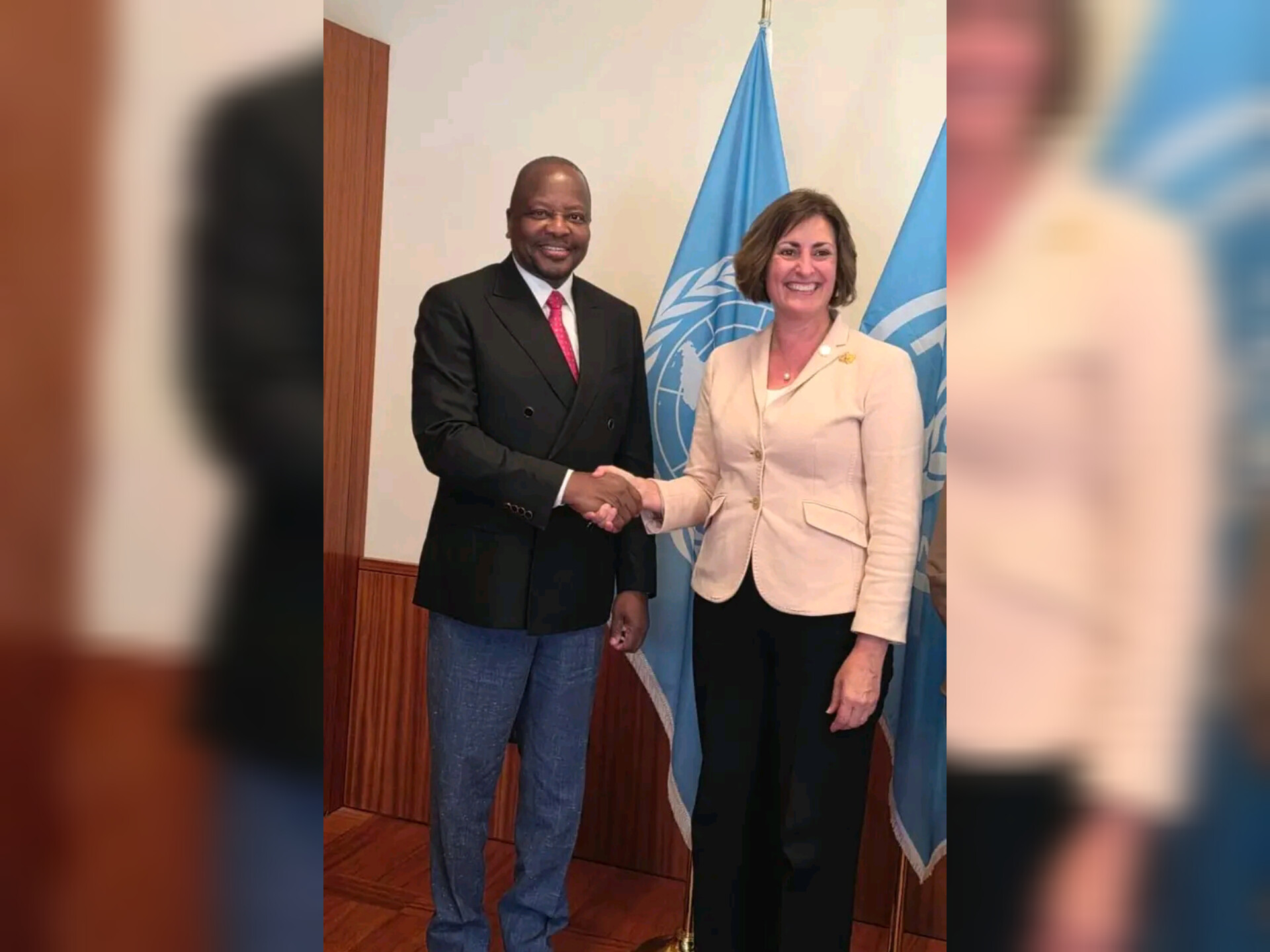 Two adults shake hands indoors, a man in a dark suit and red tie with a woman in a light blazer, standing before a United Nations flag.