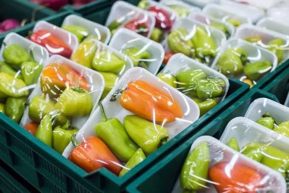 Assorted bell peppers (orange, yellow, green) packed in white foam trays, arranged in green crates at a market stall.