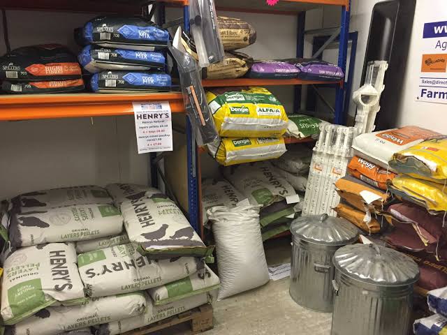 A store aisle with metal shelves stacked with large bags of Henry's animal feed and other bulk pet/farm foods; two metal bins sit in the foreground.