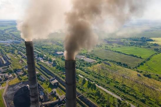 Aerial view of two tall smokestacks belching dark smoke from an industrial facility amid green fields and farmland.