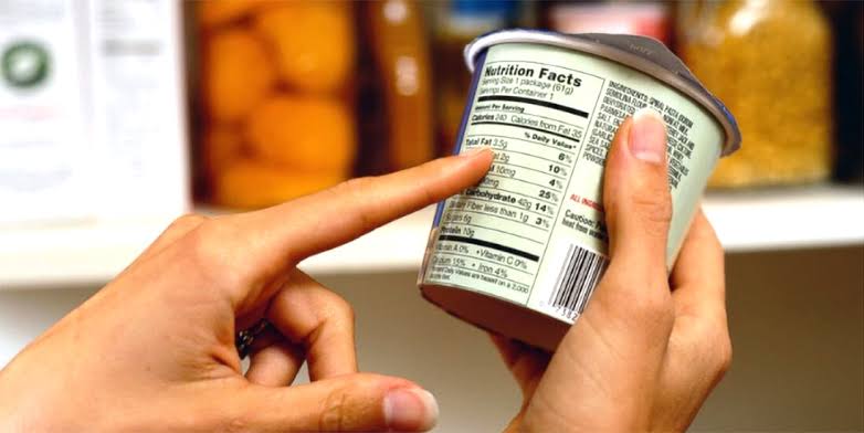 Two hands hold a yogurt cup while another points to its nutrition facts label in a grocery setting.