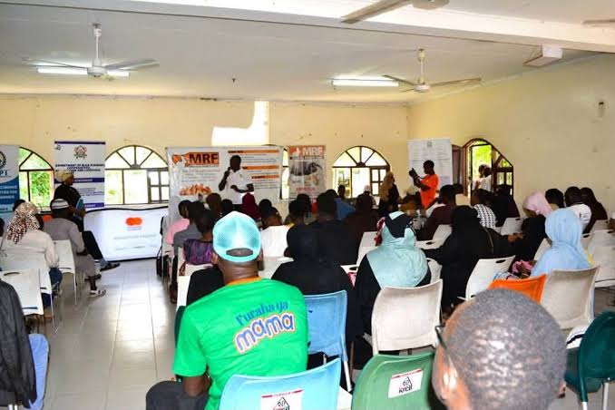 Community meeting in a bright hall with a speaker at the front and banners, attendees seated facing the stage.