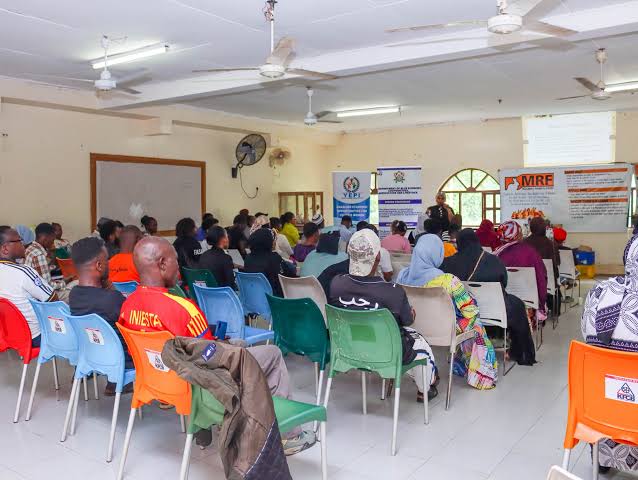 Diverse group seated in a community hall listening to a presenter at the front, with banners and a projector screen behind them.