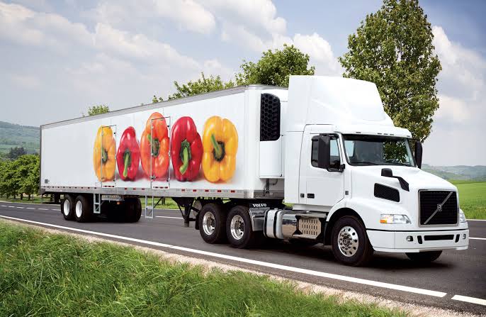 White semi-truck with a trailer displaying large bell peppers (red, yellow, orange) on the side, driving on a highway.