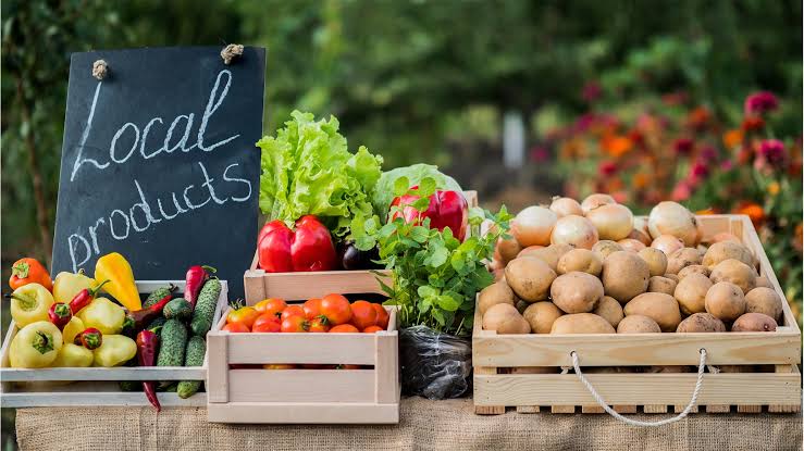 Market stall with wooden crates of fresh vegetables (peppers, cucumbers, tomatoes, lettuce) and a chalkboard reading 'Local products'.