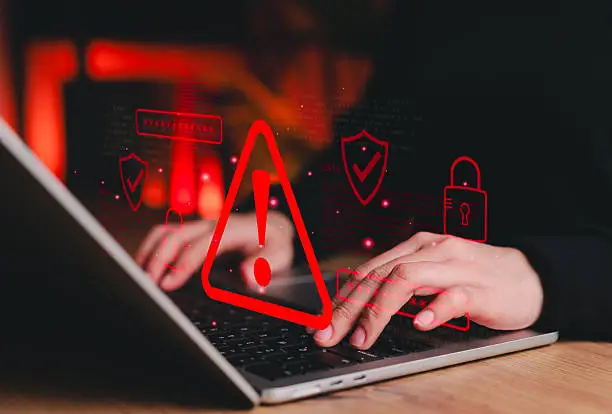 istockphoto-2240286508-612x612 - Sacco Trend Magazine Person typing on a laptop with red warning triangle and security icons floating above, conveying a cyber threat.